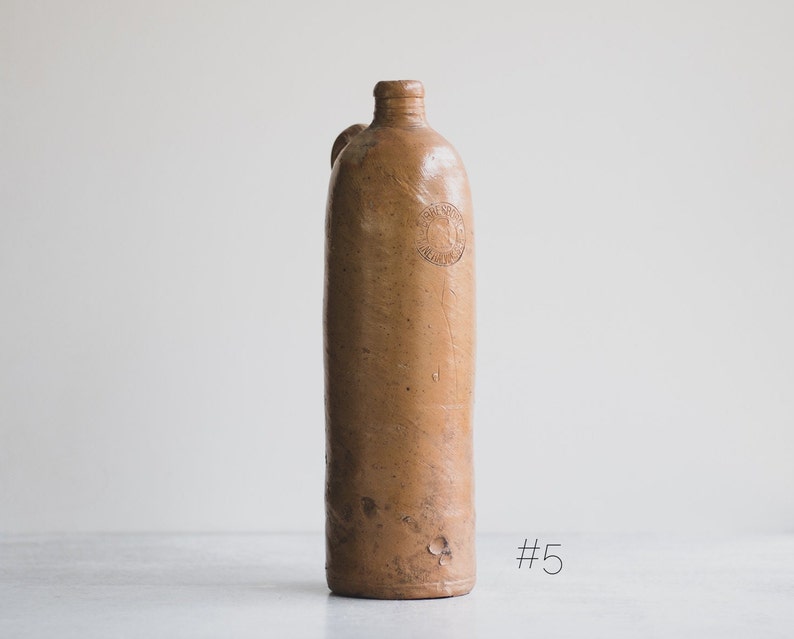 May include: An antique brown stoneware bottle with a narrow neck and a small handle. The bottle has a slightly textured surface and a circular embossed design near the top. This vintage bottle was likely used for storing liquids.