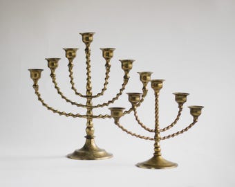 Può includere: Due antichi menorah in ottone. Ognuno ha un design a braccio curvo e attorcigliato con portacandele. I menorah sono color oro e poggiano su una superficie bianca. I menorah sono probabilmente usati per cerimonie religiose.
