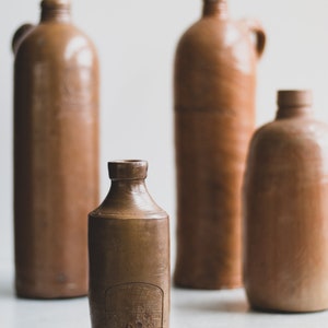 May include: Four antique brown stoneware bottles of varying sizes. The bottles have a rustic, aged appearance, with a matte finish. One bottle has a handle, and another has a label with text. The bottles are arranged against a neutral background.
