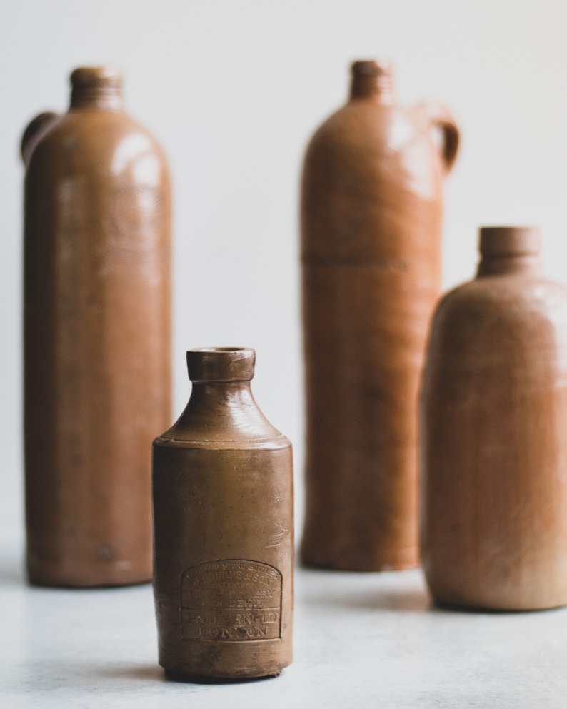 May include: Four antique brown stoneware bottles of varying sizes. The bottles have a rustic, aged appearance, with a matte finish. One bottle has a handle, and another has a label with text. The bottles are arranged against a neutral background.