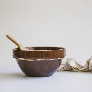 May include: A brown ceramic mixing bowl with a decorative rim and a wooden spoon inside. The bowl has a light-colored interior and a patterned exterior. A checkered cloth is draped nearby.
