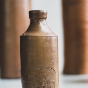 May include: A brown stoneware bottle with a cylindrical body and a narrow neck. The bottle has a label with text that is difficult to read. Two other similar bottles are blurred in the background. The bottle is likely vintage.