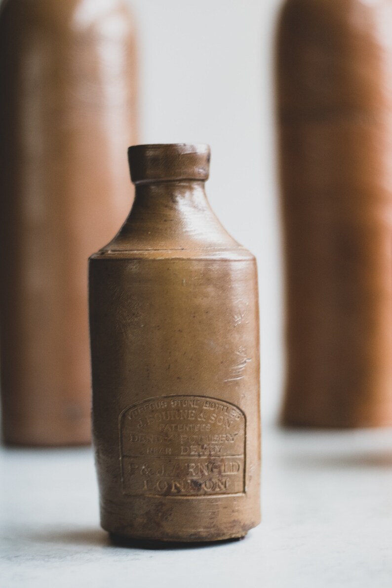 May include: A brown stoneware bottle with a cylindrical body and a narrow neck. The bottle has a label with text that is difficult to read. Two other similar bottles are blurred in the background. The bottle is likely vintage.