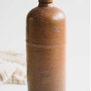 May include: A brown stoneware bottle with a rounded top and a narrow neck. The bottle has a cylindrical body and a smooth, glossy finish. The number "#6" is printed in black on the lower right side of the bottle.
