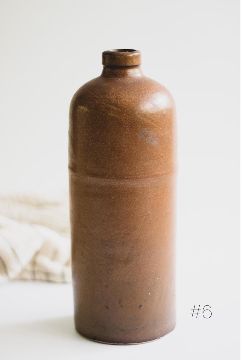 May include: A brown stoneware bottle with a rounded top and a narrow neck. The bottle has a cylindrical body and a smooth, glossy finish. The number "#6" is printed in black on the lower right side of the bottle.
