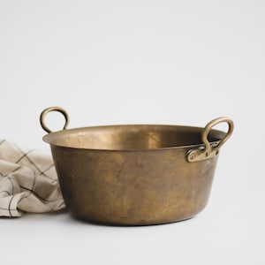 May include: A vintage brass bowl with two loop handles. The bowl has a rounded shape and a slightly aged appearance. A folded, cream-colored cloth with a black grid pattern is to the left of the bowl.