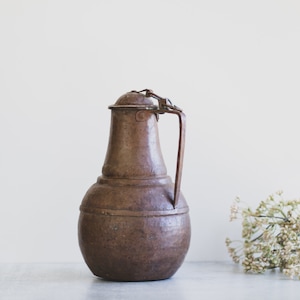 May include: A vintage copper pitcher with a hinged lid and a handle. The pitcher has a rustic brown color and a slightly rounded shape.