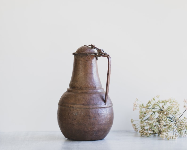 May include: A vintage copper pitcher with a hinged lid and a handle. The pitcher has a rustic brown color and a slightly rounded shape.