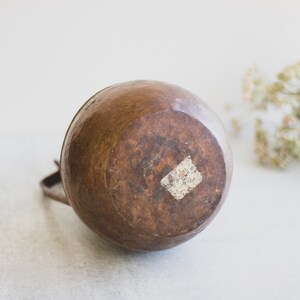 May include: A copper pitcher with a rounded body and a handle. The pitcher has a worn, aged look and a small white sticker on the bottom.