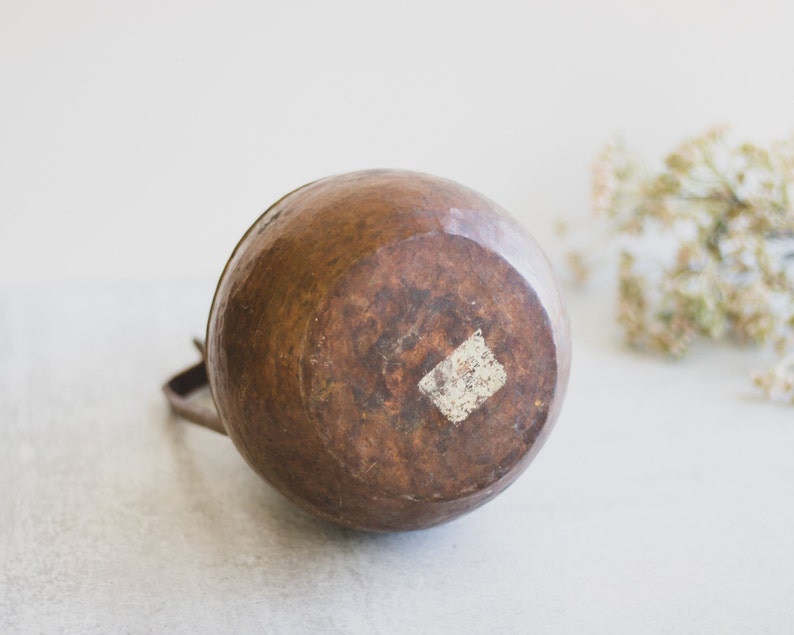 May include: A copper pitcher with a rounded body and a handle. The pitcher has a worn, aged look and a small white sticker on the bottom.