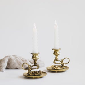 May include: Two antique brass candle holders with lit white candles. The candle holders have a decorative design with a circular base and a handle. A beige cloth is in the background. The candle holders are a vintage home decor item.
