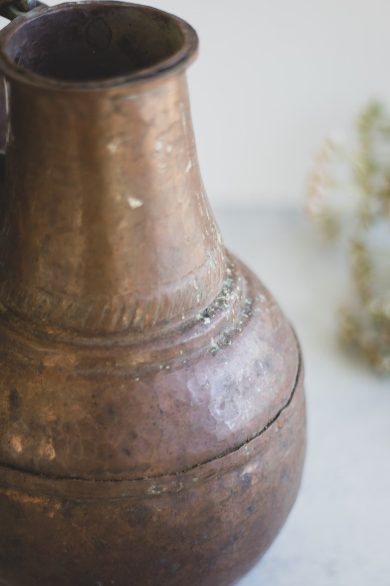 May include: A vintage copper pitcher with a worn, aged patina. The pitcher has a round body and a narrow neck with a handle.