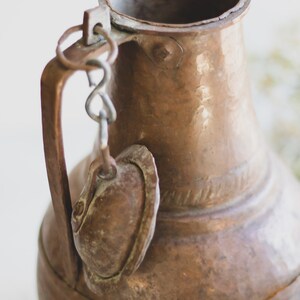 May include: A copper pitcher with a handle and a small lid attached by a chain. The pitcher has a hammered finish and a worn patina.