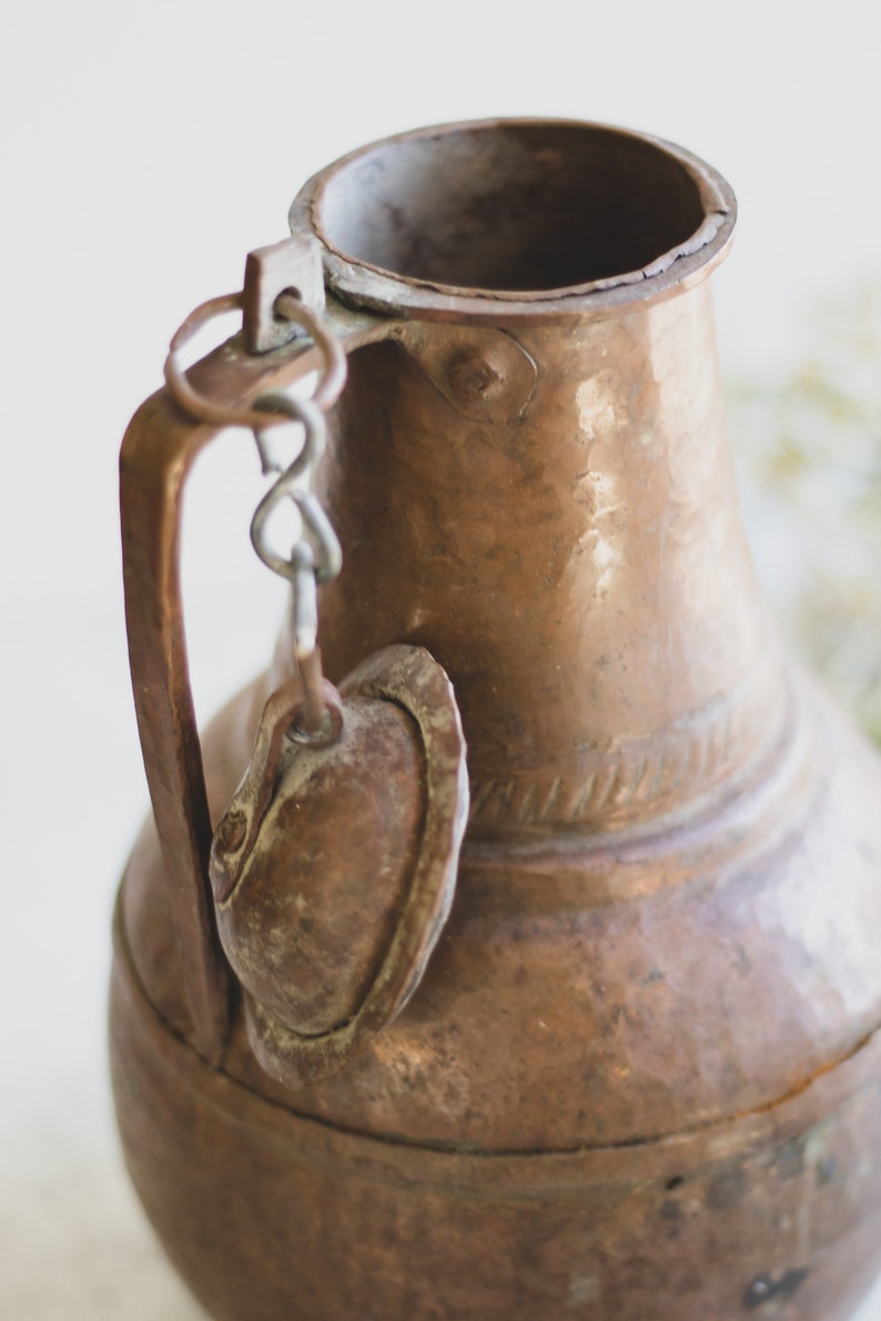 May include: A copper pitcher with a handle and a small lid attached by a chain. The pitcher has a hammered finish and a worn patina.