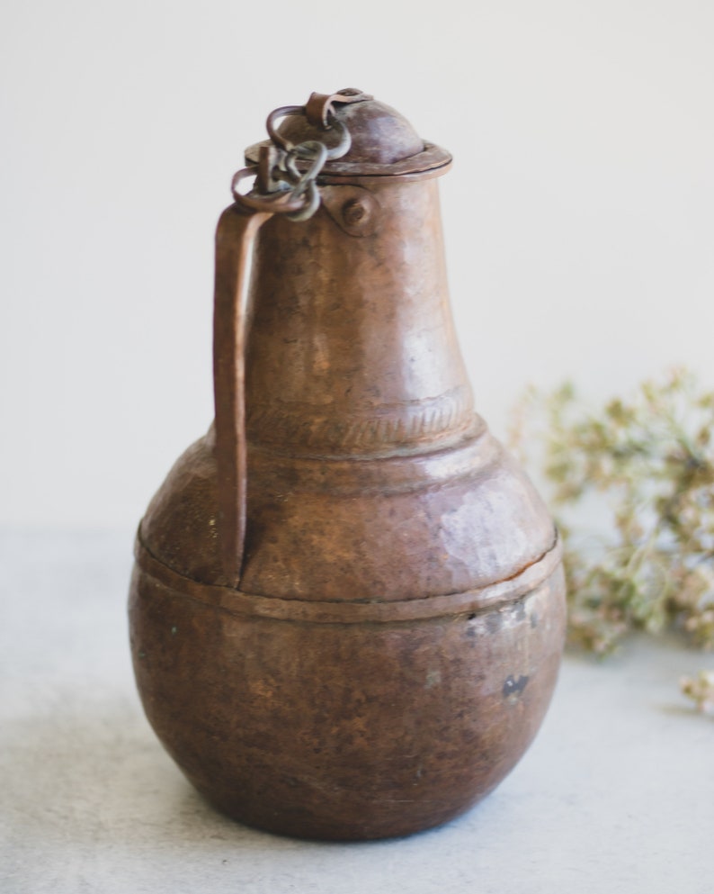 May include: A vintage copper pitcher with a hammered finish and a chain handle. The pitcher has a rounded body and a narrow neck.
