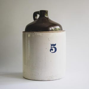 May include: A vintage stoneware jug with a dark brown glazed top and handle. The off-white body features a large blue number "5". The jug is cylindrical with a rounded shoulder and a narrow neck.