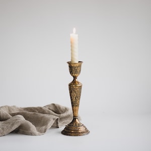 Puede incluir: Un candelabro de latón ornamentado sostiene una vela en espiral blanca encendida. El candelabro presenta intrincados diseños florales y un diseño alto y esbelto. Un paño de lino beige está drapeado cerca, creando un fondo suave y neutro.