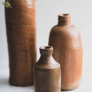 May include: Three antique brown stoneware bottles of varying sizes. The tallest bottle has a handle. The smallest bottle has a label with text. The bottles are arranged in front of a white background.