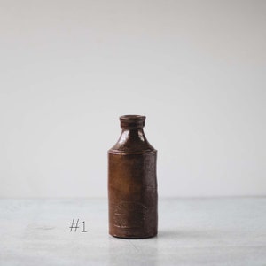 May include: A brown stoneware bottle with a narrow neck and a cylindrical body. The bottle has a dark, glossy finish and a small, embossed design near the base. The number "#1" is written in black ink on the surface.