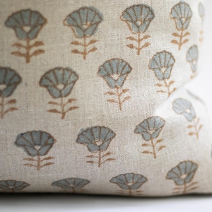 May include: A beige linen fabric cushion cover featuring a repeating pattern of stylised flowers. Each flower has light blue petals and a brown stem and leaves, printed in a consistent grid-like arrangement across the textured textile surface.