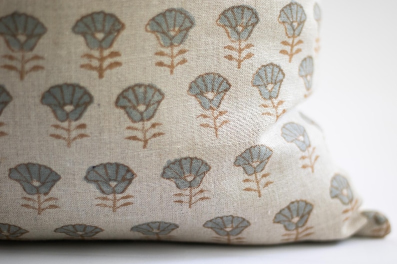 May include: A beige linen fabric cushion cover featuring a repeating pattern of stylised flowers. Each flower has light blue petals and a brown stem and leaves, printed in a consistent grid-like arrangement across the textured textile surface.