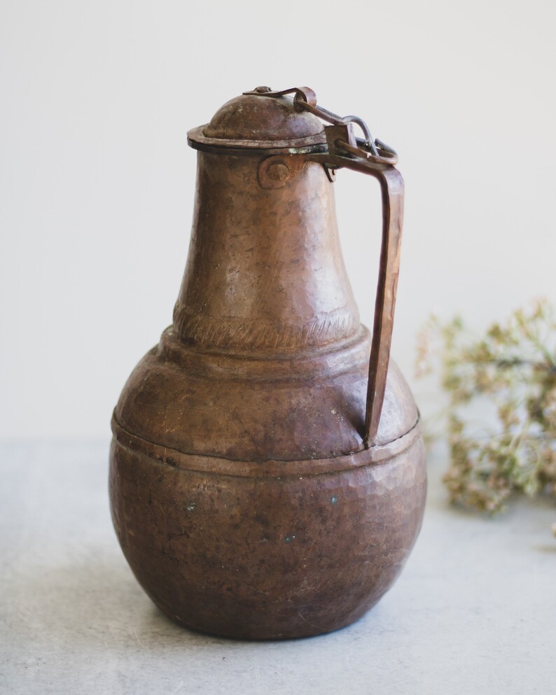 May include: A vintage copper pitcher with a hinged lid and a handle. The pitcher has a rounded body and a slightly flared spout. The copper has a patina of age and wear.