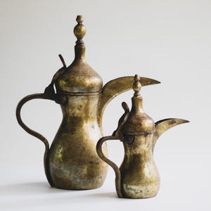 May include: Two antique brass coffee pots with ornate designs. The pots have curved spouts, handles, and lids with decorative finials. The larger pot is approximately 12 inches tall, and the smaller pot is about 8 inches tall. The brass has a weathered, aged appearance.