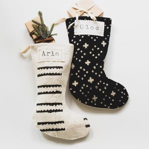 Op de afbeelding: Twee kerstsokken met gepersonaliseerde naamlabels. De ene sok is gebroken wit met zwarte horizontale zigzagstrepen en de naam Arlo. De andere is zwart met een wit kruispatroon en de naam Cloe. Beide sokken zijn voorzien van kleine cadeauverpakkingen.