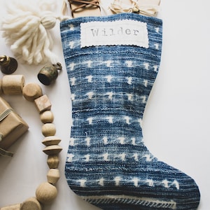 May include: A blue and white patterned fabric Christmas stocking featuring a white rectangular patch with the name Wilder printed in black text. The stocking is decorated with a large white yarn tassel and is surrounded by wooden beads and gift boxes.