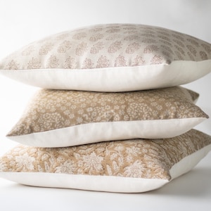 May include: A stack of three square throw cushions. The top cushion is beige with a small brown floral pattern. The middle cushion is tan with a dense white floral print. The bottom cushion is tan with a large white floral pattern on the front.