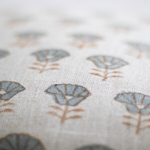 May include: A light beige fabric cushion cover featuring a repeating pattern of small, stylised flowers. Each flower has light blue petals and a brown stem and leaves. The fabric has a visible woven texture, and the focus is on the centre.