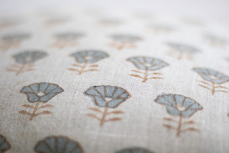 May include: A light beige fabric cushion cover featuring a repeating pattern of small, stylised flowers. Each flower has light blue petals and a brown stem and leaves. The fabric has a visible woven texture, and the focus is on the centre.