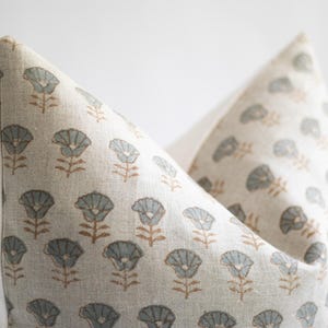 May include: A square throw cushion cover made of light beige fabric featuring a repeating pattern of small, stylised flowers. Each flower has a muted blue-grey petal section and a thin brown stem, printed in a consistent grid across the front surface.