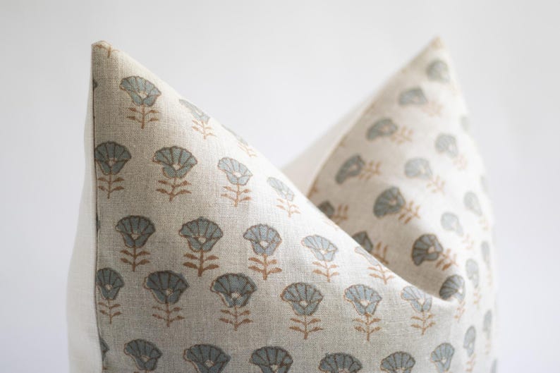 May include: A square throw cushion cover made of light beige fabric featuring a repeating pattern of small, stylised flowers. Each flower has a muted blue-grey petal section and a thin brown stem, printed in a consistent grid across the front surface.