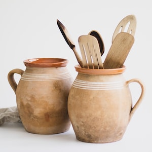 May include: Two rustic, earthenware pitchers with handles. One pitcher holds wooden kitchen utensils, including a spoon and slotted spatulas. The pitchers are tan with a brown rim and white stripes. The background is white.