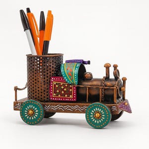 Puede incluir: Un portalápices decorativo con forma de tren antiguo. El tren de metal marrón presenta un portabolígrafos perforado, detalles coloridos y ruedas verdes. Hay bolígrafos naranjas, negros y blancos dentro del portalápices.