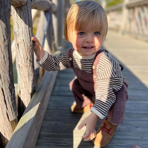 May include: A young child wearing a striped long-sleeve shirt and corduroy overalls. The child is wearing brown shoes with red accents and is crouching on a wooden bridge. The background is blurred, with sunlight illuminating the scene.