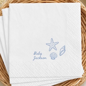 Pode incluir: Uma pilha de guardanapos de papel branco com um design impresso em azul de uma estrela-do-mar, uma concha e um búzio, com o texto Baby Jackson impresso em letra cursiva azul. Os guardanapos têm uma borda texturizada com padrão de losangos.