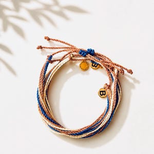 May include: A stack of woven bracelets in shades of brown, blue, and cream. Each bracelet features a gold charm with the letter "B". The bracelets are tied with brown and blue threads.