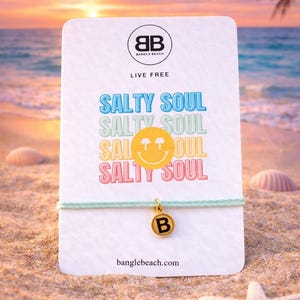 Può includere: Una carta bianca con la scritta "Salty Soul" a lettere colorate, una faccina sorridente e un braccialetto con ciondolo dorato con la lettera "B". La carta è su una spiaggia sabbiosa con un tramonto sullo sfondo. Il marchio è "Bangle Beach".