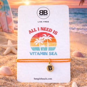 Può includere: Un braccialetto con cordino arancione con un ciondolo dorato con la lettera "B" è appeso davanti a una carta con la scritta "ALL I NEED IS VITAMIN SEA". La carta ha una grafica a tema spiaggia e le parole "LIVE FREE" e "banglebeach.com".