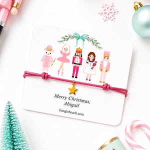 Pode incluir: Uma pulseira de cordão rosa com um pingente de estrela dourada num cartão branco com ilustrações de um rato, uma bailarina, um quebra-nozes, uma menina e outro quebra-nozes. O cartão inclui o texto Merry Christmas, Abigail e banglebeach.com.