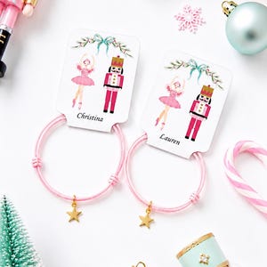 Peut inclure: Deux élastiques à cheveux roses avec des breloques en forme d'étoile dorée, fixés sur des cartes de présentation blanches illustrées d'une ballerine et d'un casse-noisette. Les cartes sont personnalisées avec les prénoms Christina et Lauren.