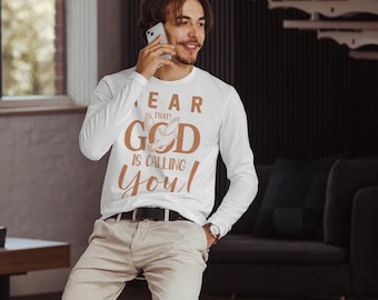 Peut inclure: T-shirt blanc à manches longues avec l'inscription "HEAR THAT? GOD IS CALLING YOU!" en marron. Le t-shirt présente un motif de colombe. La personne porte une ceinture noire et un pantalon beige.