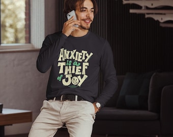 Peut inclure: Un t-shirt bleu marine à manches longues avec l'inscription "Anxiety is a Thief of Joy" dans une police fantaisiste. Le t-shirt est associé à un pantalon clair et une ceinture noire. La personne sur l'image est au téléphone.