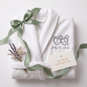 May include: A white plush robe featuring grey embroidery of two hearts with the letters S and M, and the date July 25, 2026. A sage green ribbon is tied around the robe, accompanied by a small bouquet of dried lavender and a card reading For S & M on their Special Day.