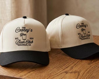 Op de afbeelding: Twee baseballpetten met gebroken witte kronen en zwarte kleppen. Elke pet is voorzien van zwarte tekst met de tekst The Cooney's Bach Club, EST. 2025, Savannah, GA, vergezeld van een afbeelding van een drankje met een schijfje limoen.