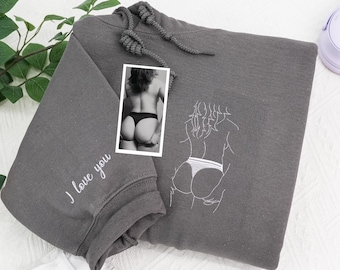 May include: A charcoal gray hooded sweatshirt featuring a white embroidered line art illustration of a person from behind wearing underwear. The sleeve includes the embroidered text I love you. A small rectangular photograph shows the same subject.