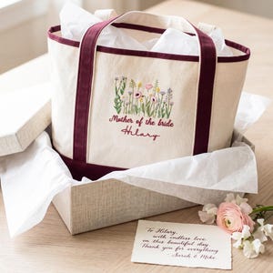 May include: A cream-colored tote bag with burgundy trim and handles, embroidered with wildflowers and the text "Mother of the bride Hilary." The bag is nestled in a gift box with tissue paper, accompanied by a handwritten card.