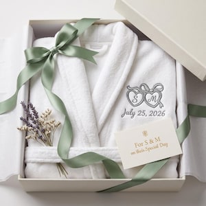 May include: A white plush bathrobe folded in a gift box, tied with a sage green ribbon. The robe features grey embroidery of two hearts with the letters S and M and the date July 25, 2026. A small card reads For S & M on their Special Day.